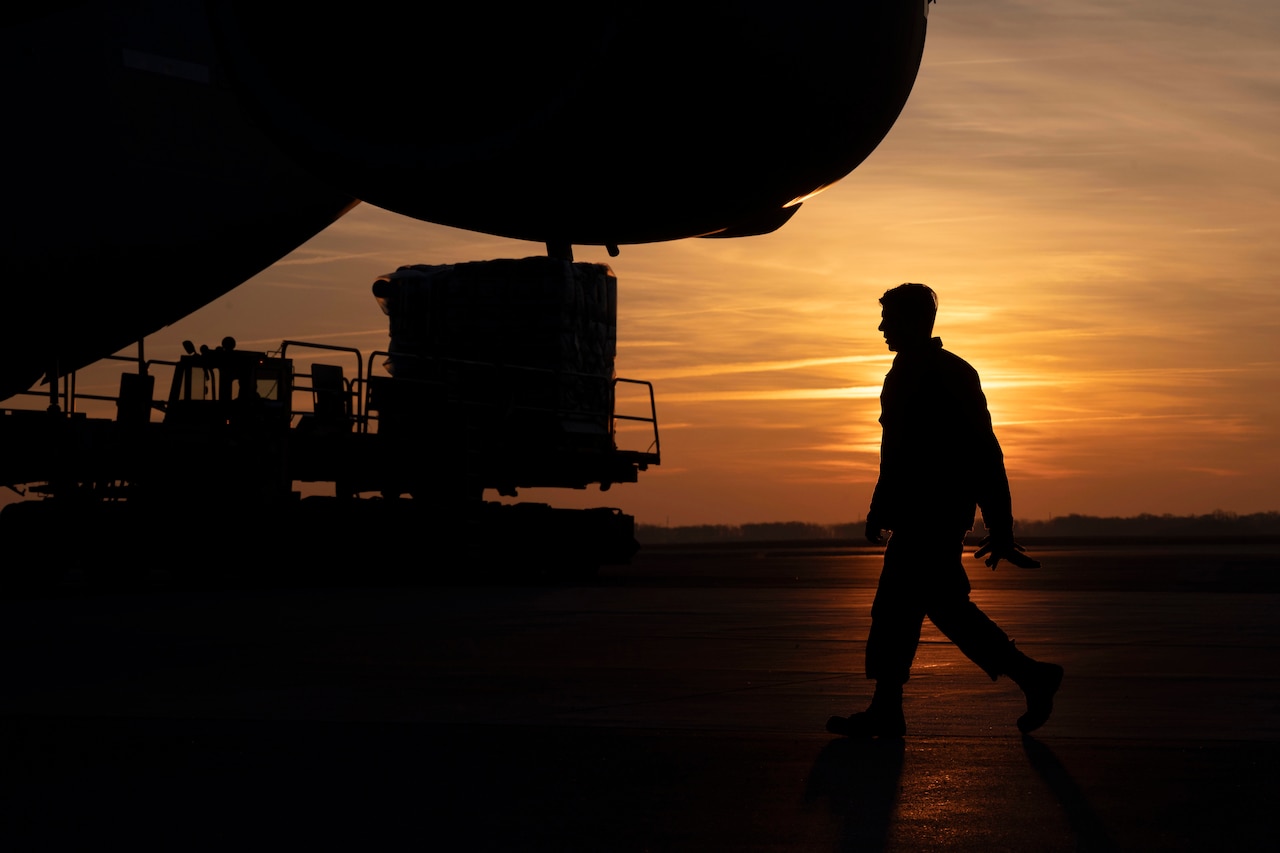 An airman in silhouette walks near an aircraft on flight line against orange sky.