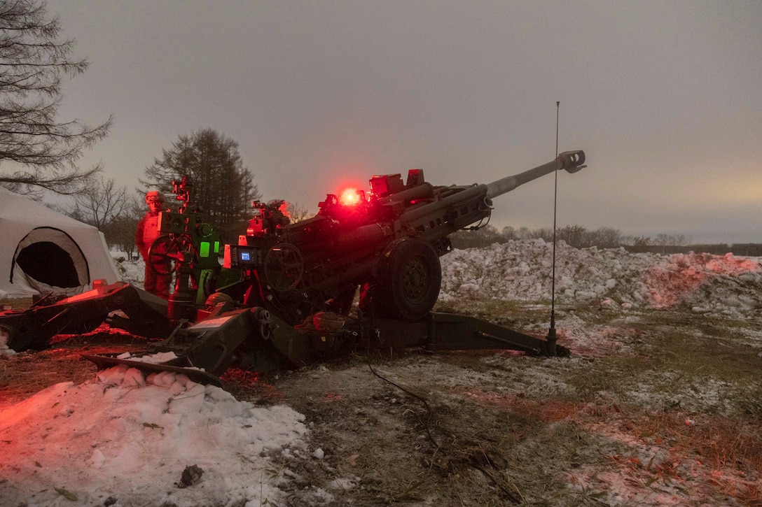 A Marine stands near a weapon at a tent in the snow illuminated by a red light.