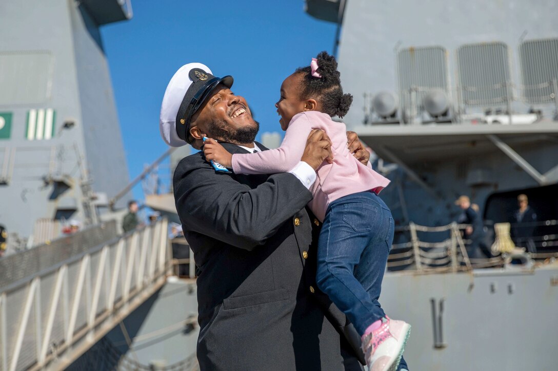 A sailor smiles while picking up his daughter near a ship.