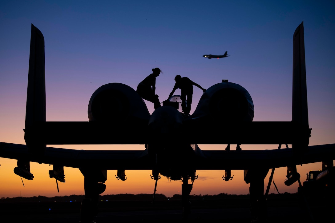 Two airmen shown in silhouette work on top of a parked aircraft.