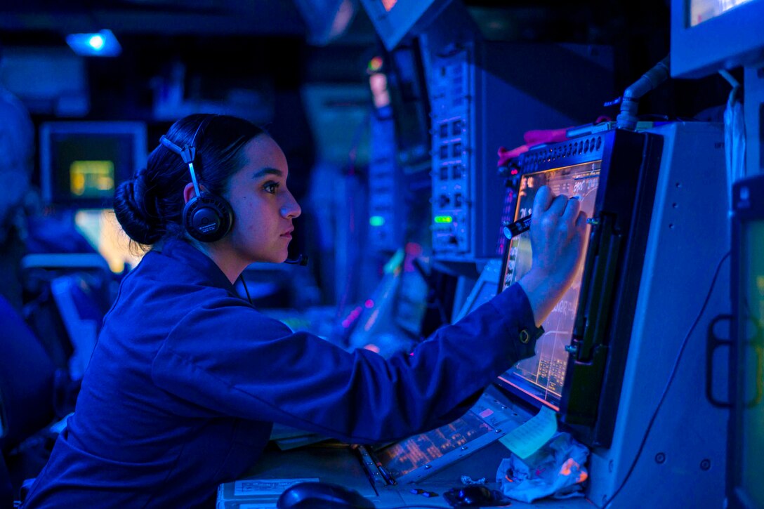 A sailor illuminated by blue light sits in front of digital monitors.