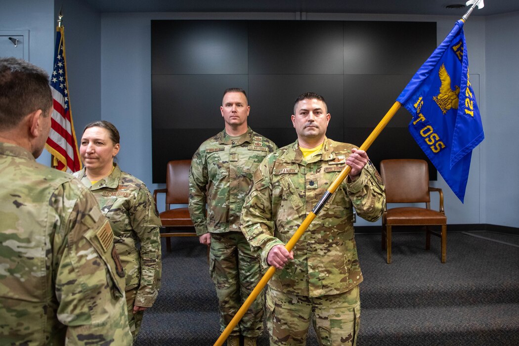 Four uniformed servicemembers stand in ceremonial fashion while one male servicemember holds flag in presentation manner.