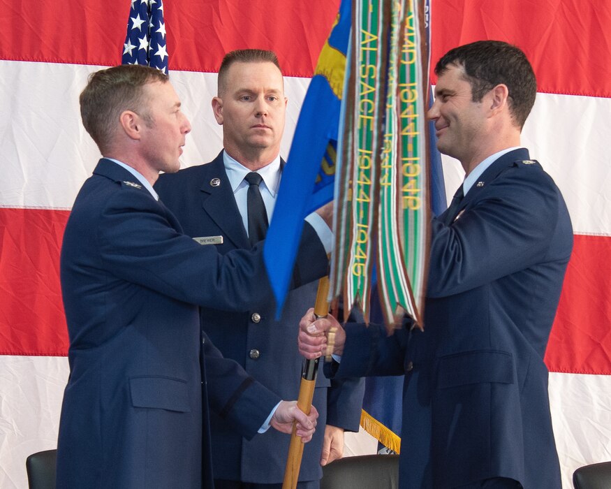 Group commander and incoming squadron commander facing each other while each holds the unit flag, group commander is speaking to squadron commander who is smiling. The first sergeant stands behind, looking on.