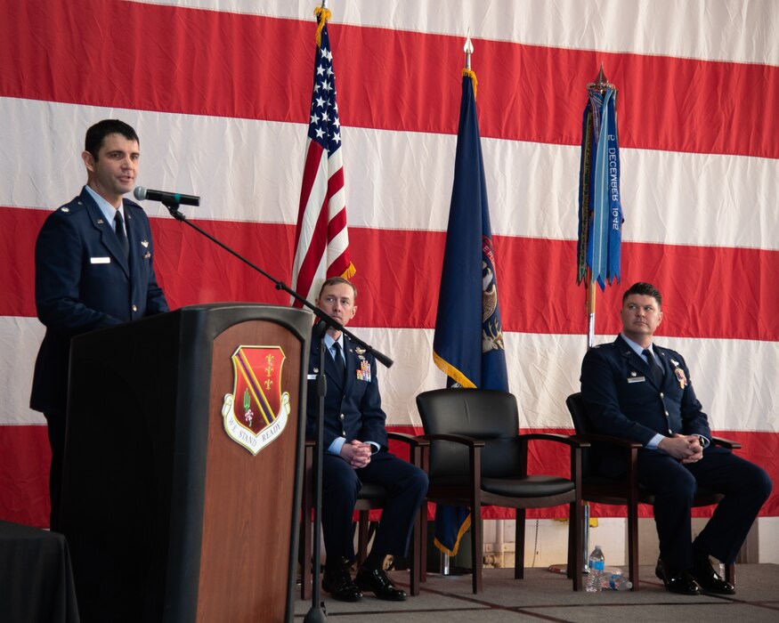 Three male service members in dress uniform on stage. One stands at lectern and the two others are seated, listening.