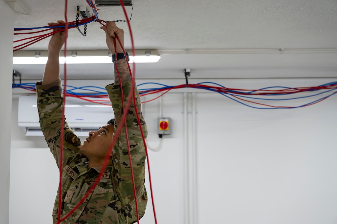 An airman reaches to adjust red and blue cables hanging from a ceiling.