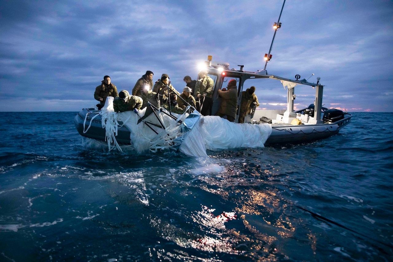 Sailors pull white canvas-type material aboard a small waterborne vessel against a dark blue sky.