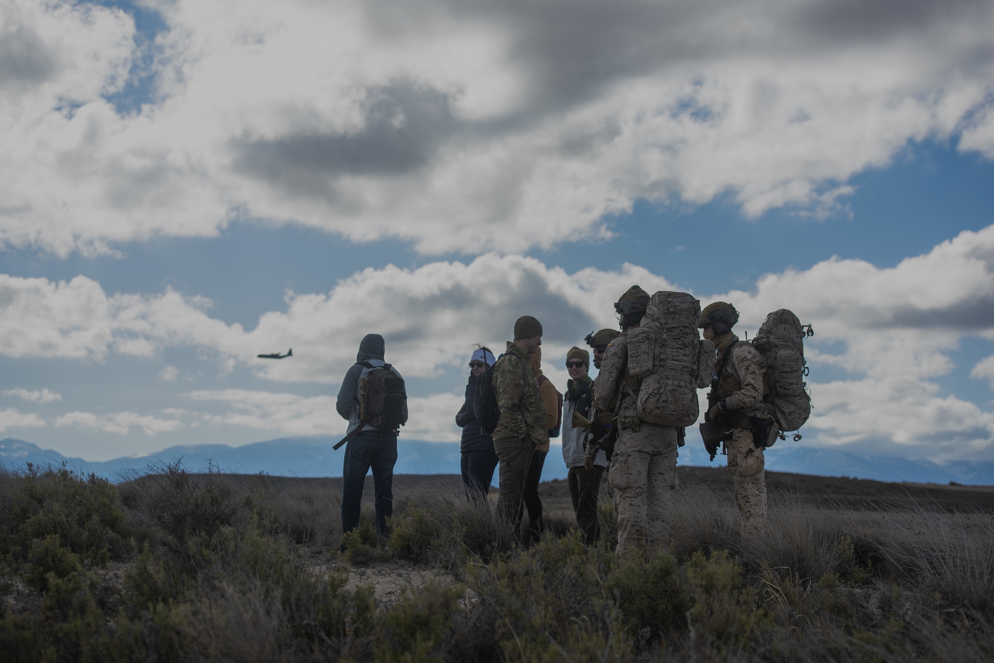 Seven people stand on a mountain with a C-130J flying overhead