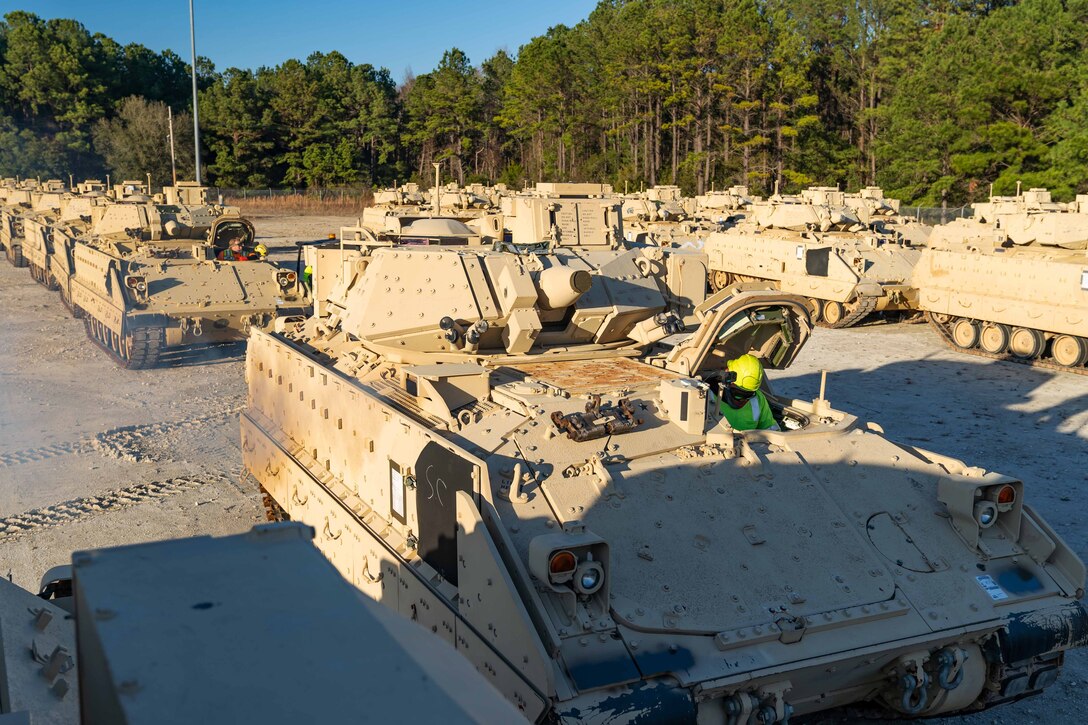 A soldier sits in a tank among rows of tanks in a sandy area.