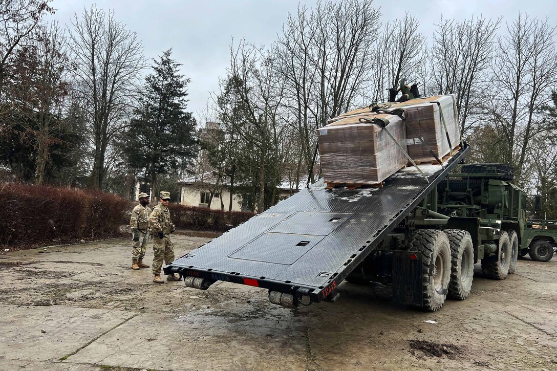 Two soldiers watch as large packages are unloaded from a flatbed vehicle.