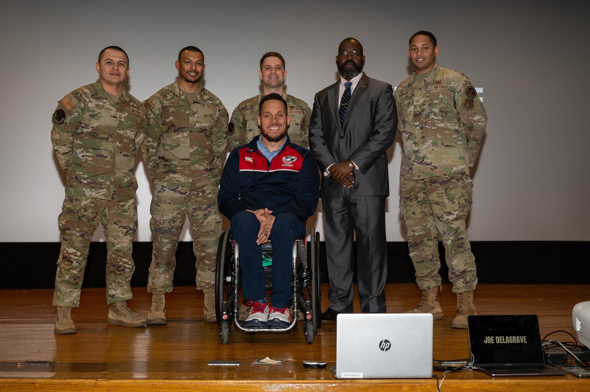 A man in a wheelchair faces the camera with five military men standing behind him