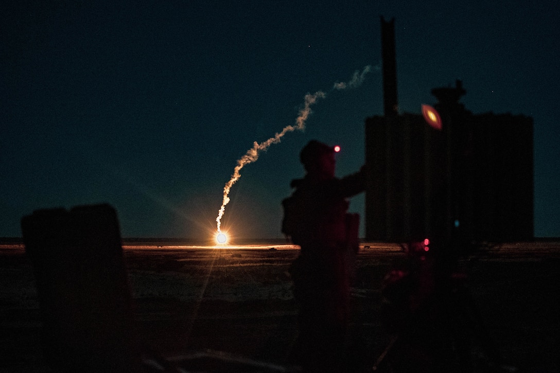 A flare creates a trail in the distance behind a Marine operating equipment in the dark.