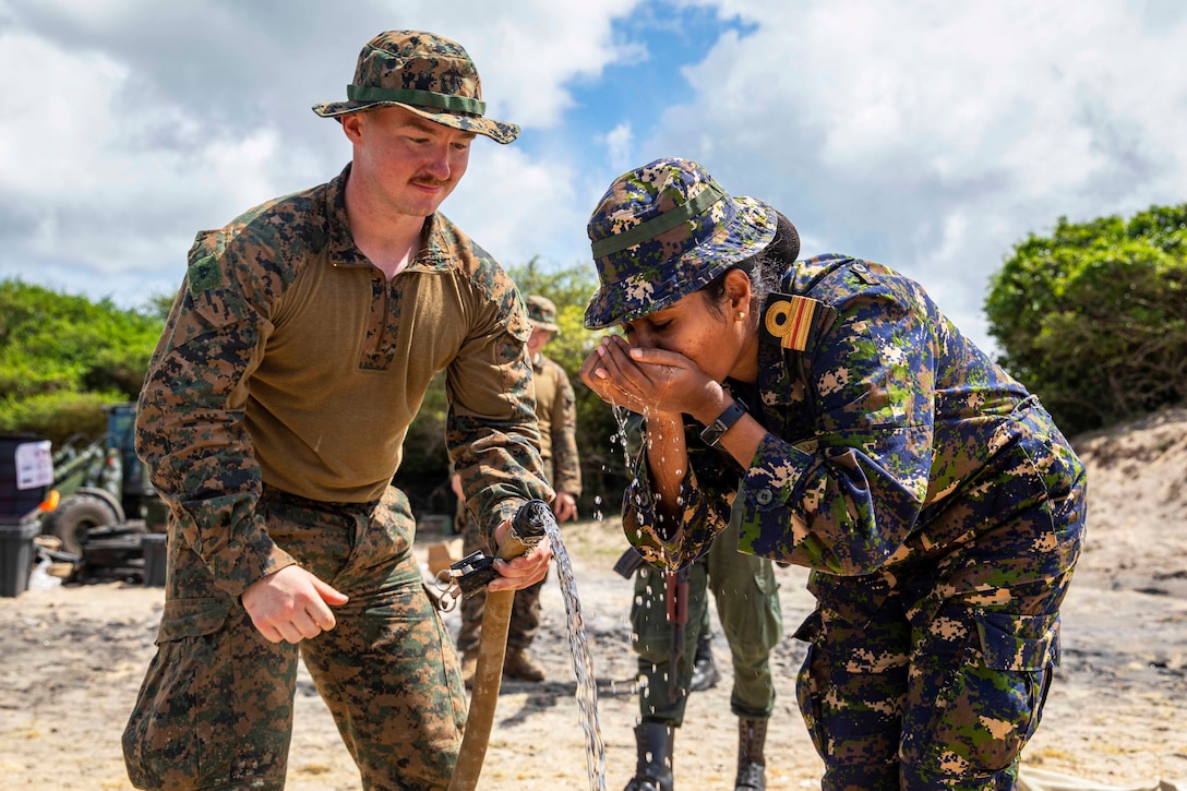 A Marine holds a hose and watches as a foreign service member drinks water.