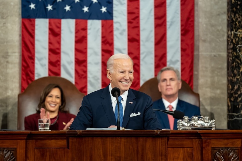 President Joe Biden stands at lectern in front of U.S. flag  and seated vice president and House speaker.