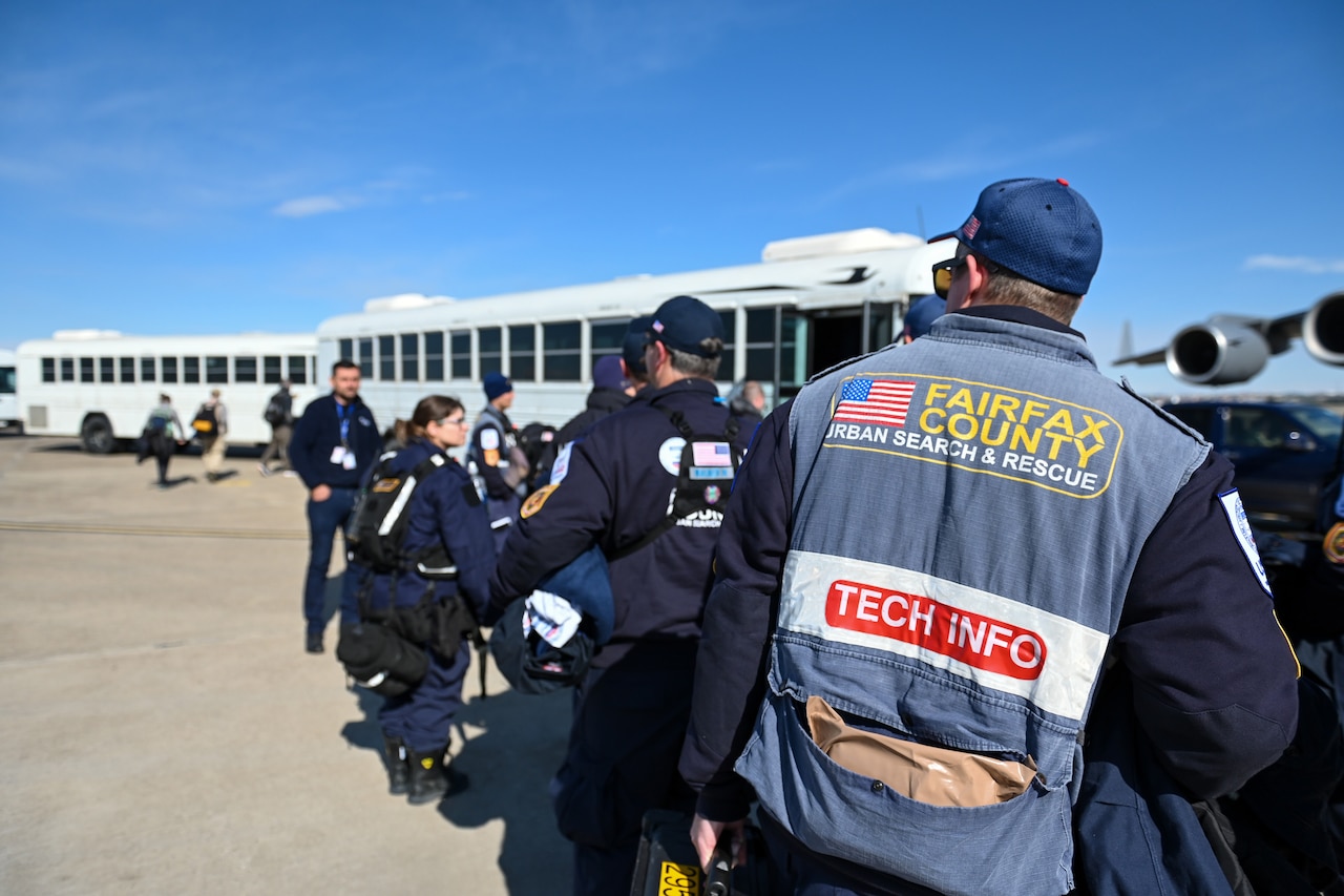 Lines of people walk toward awaiting buses. One of the people is wearing a vest that indicates he is from Fairfax County, Va., search and rescue team.