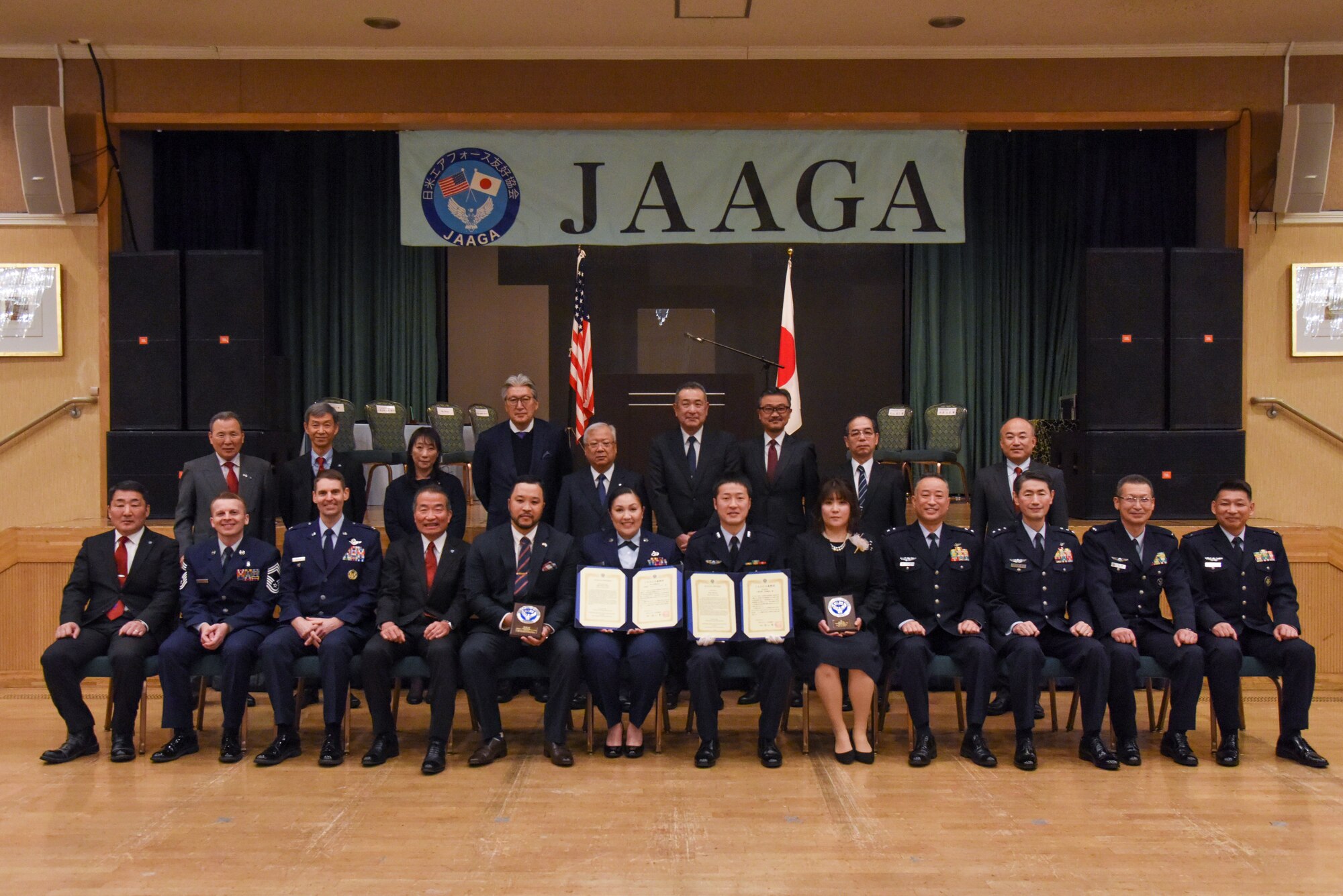 Military members and civilians pose for a group photo in front of a stage with a banner with "JAAGA" hanging above their heads.