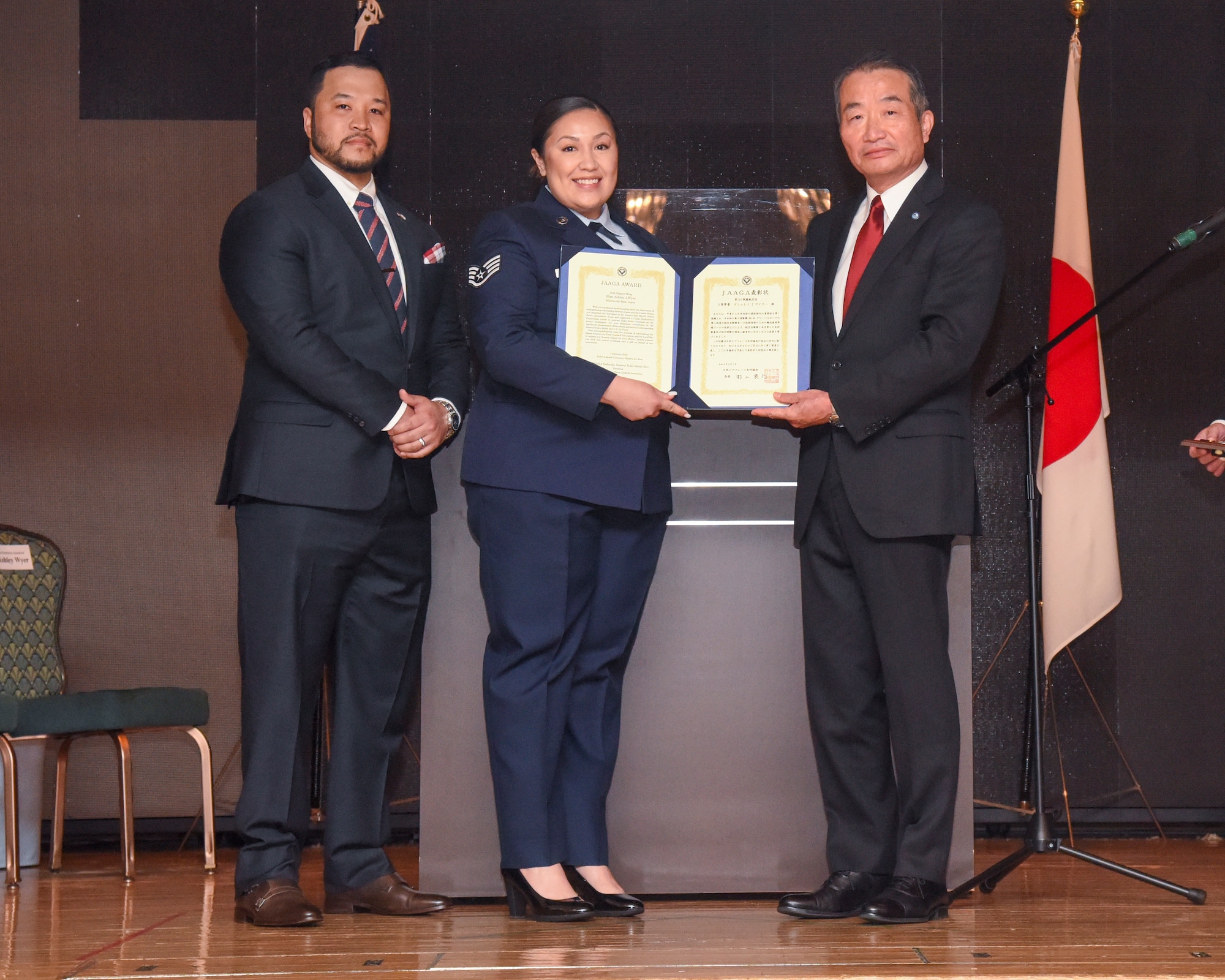 Three people stand on a stage posing for a photo with a certificate being presented.