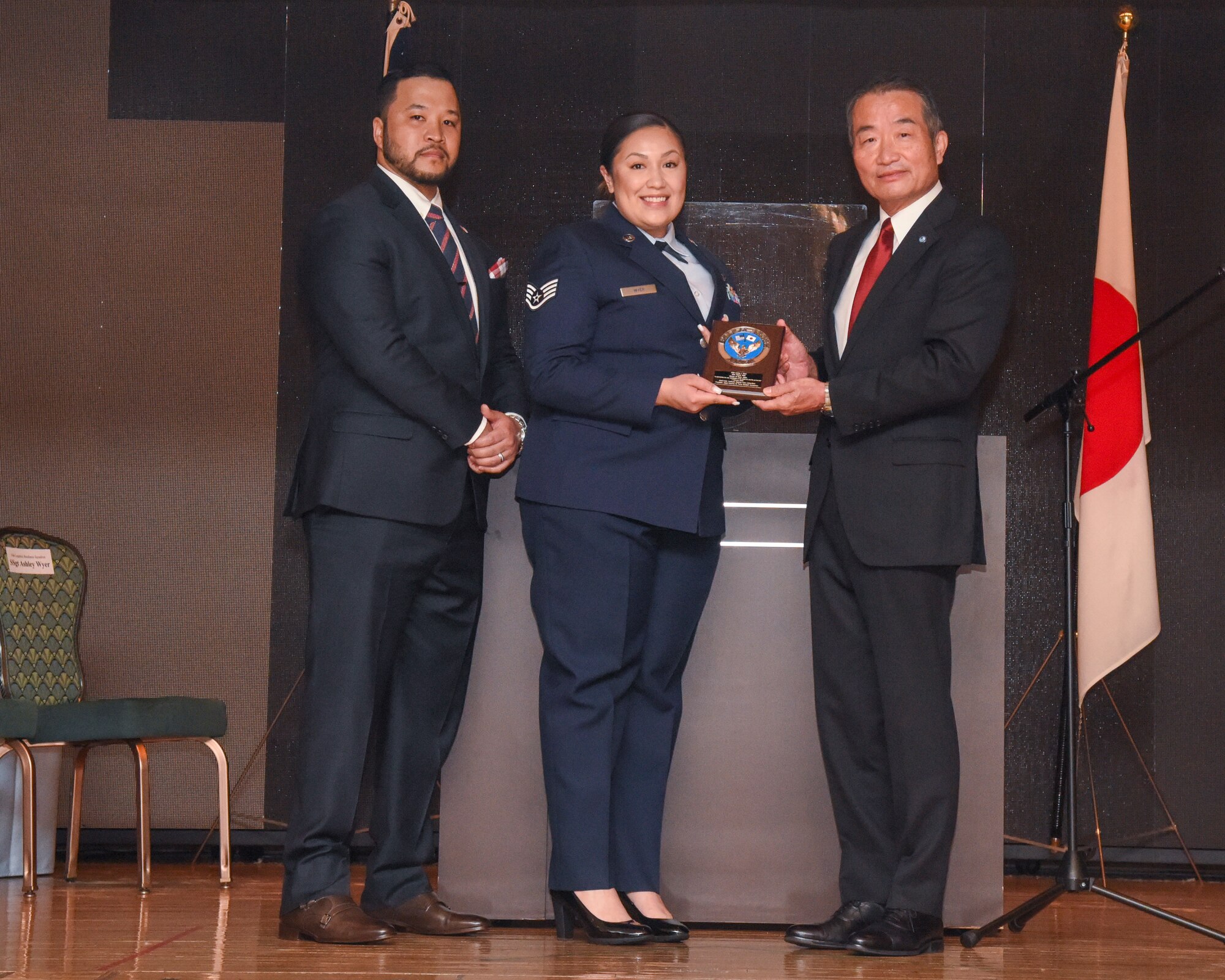 Three people stand on a stage posing for a photo with a plaque being presented.