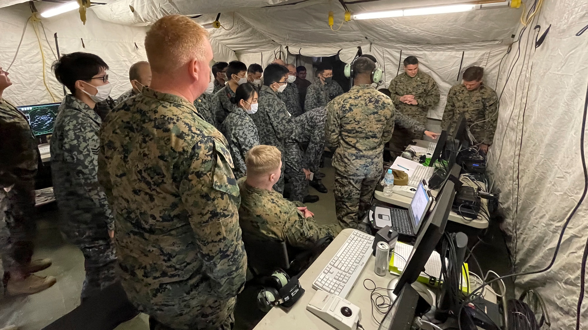 20 U.S. Marines and Japanese air force members crowd around a monitor in a large tent