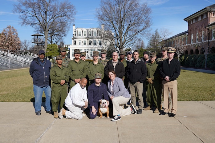 Twenty people, a combination of marines, sailors, civilians and one bulldog posing for a group photo on a grassy parade ground.