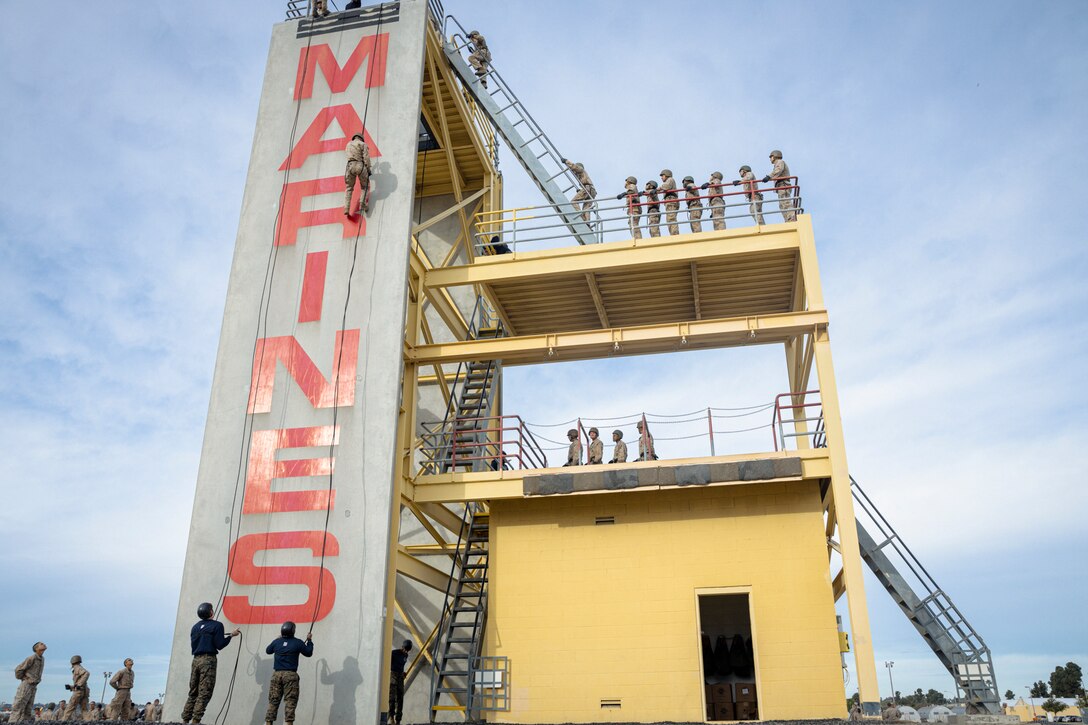 A Marine rappels down a tower as fellow Marines stand in line.