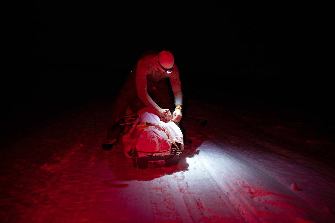 An airman opens a sled on the snow in the dark illuminated by a flashlight.
