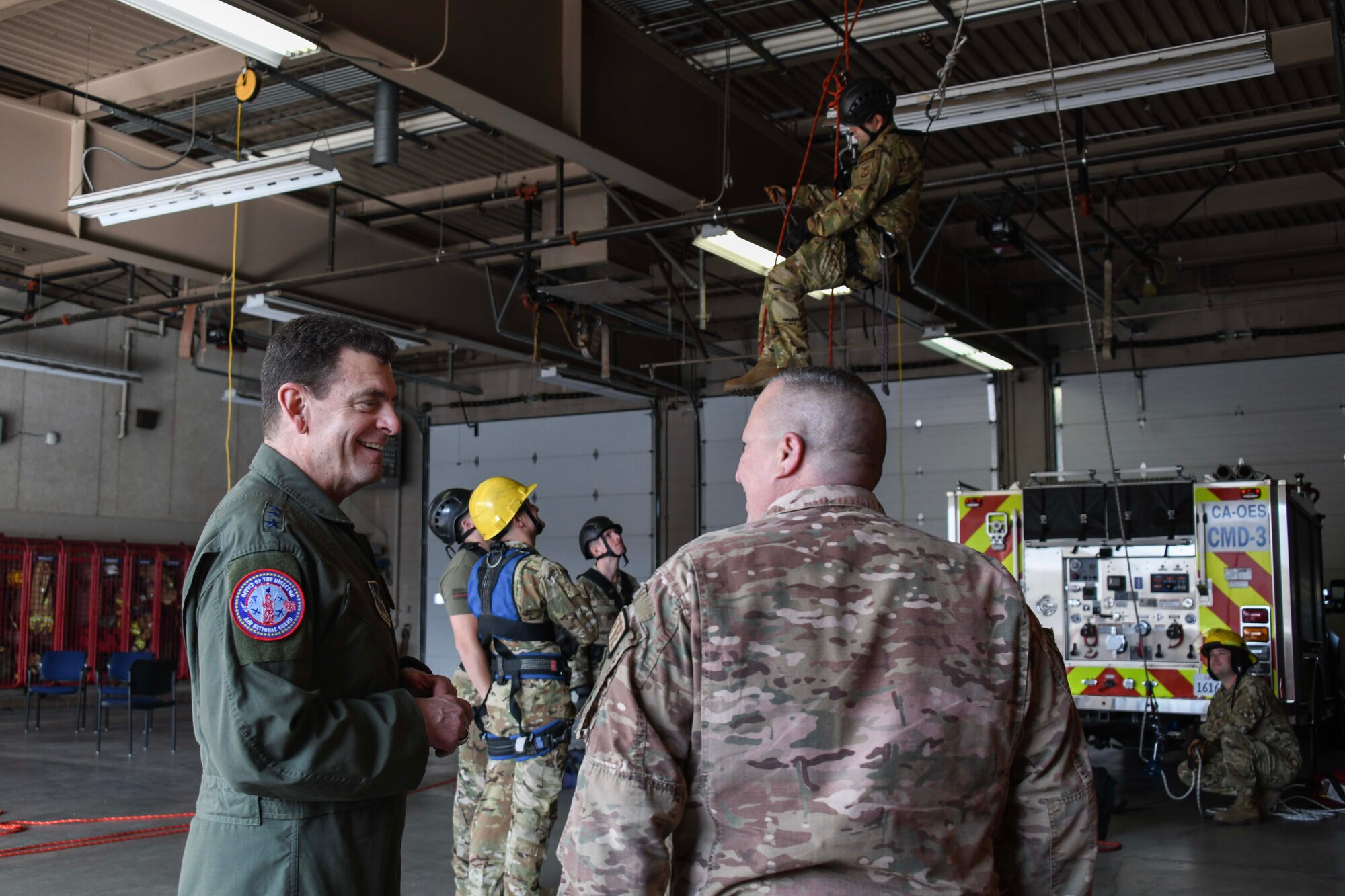 In the foreground, on the left, a man in a green military flight suit speaks to a man in a military camouflage uniform in front of firefighters doing a hanging drill