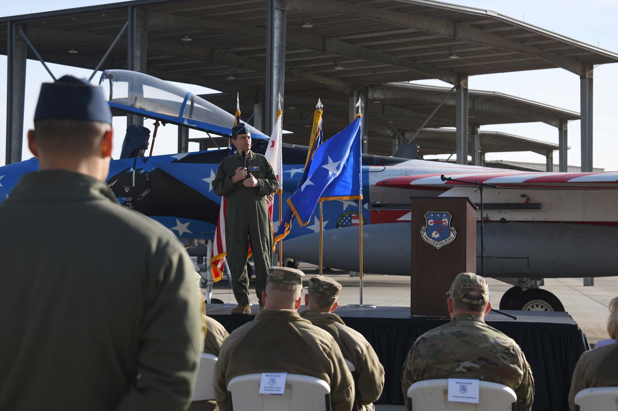 A man wearing a green military flightsuit uniform stands on a stage in front of a jet aircraft painted red, white and blue