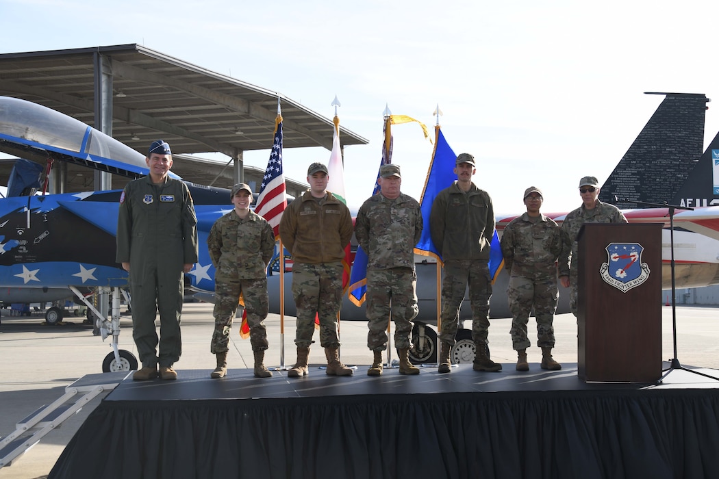 Seven military personnel wearing military camouflage uniforms stand on a stage in front of a jet aircrafted painted red white and blue