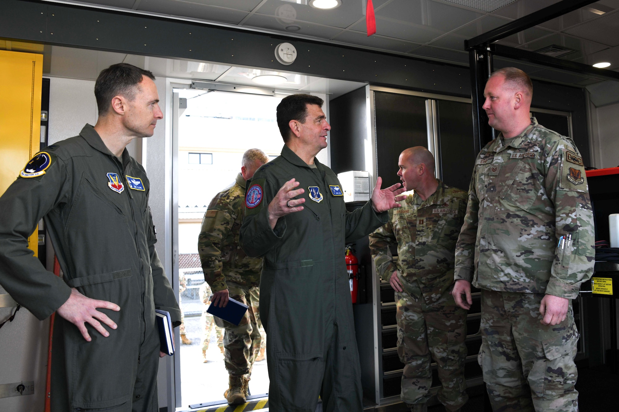 two men on the left in green military flightsuit uniforms speak to a man on the right wearing a military camouflage uniform