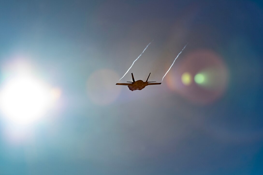 A military aircraft flies with sun rays behind it.