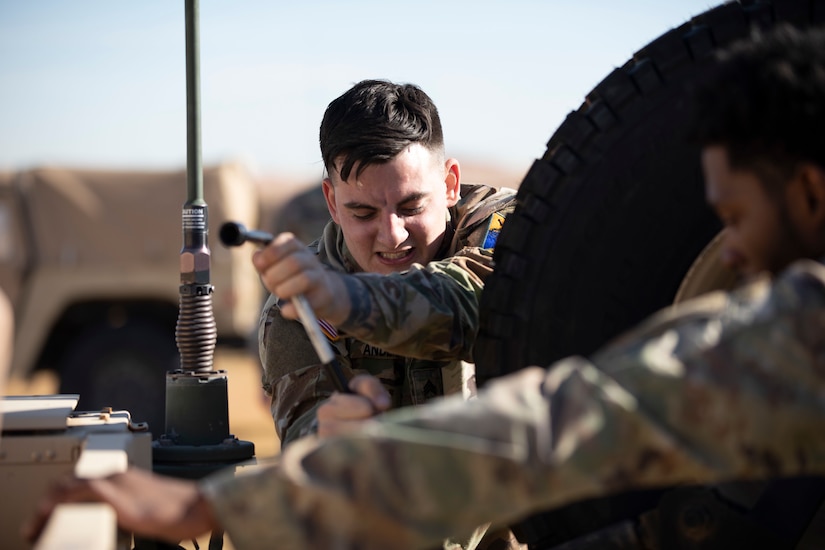 A man in military uniform puts a spare tire into a military vehicle.