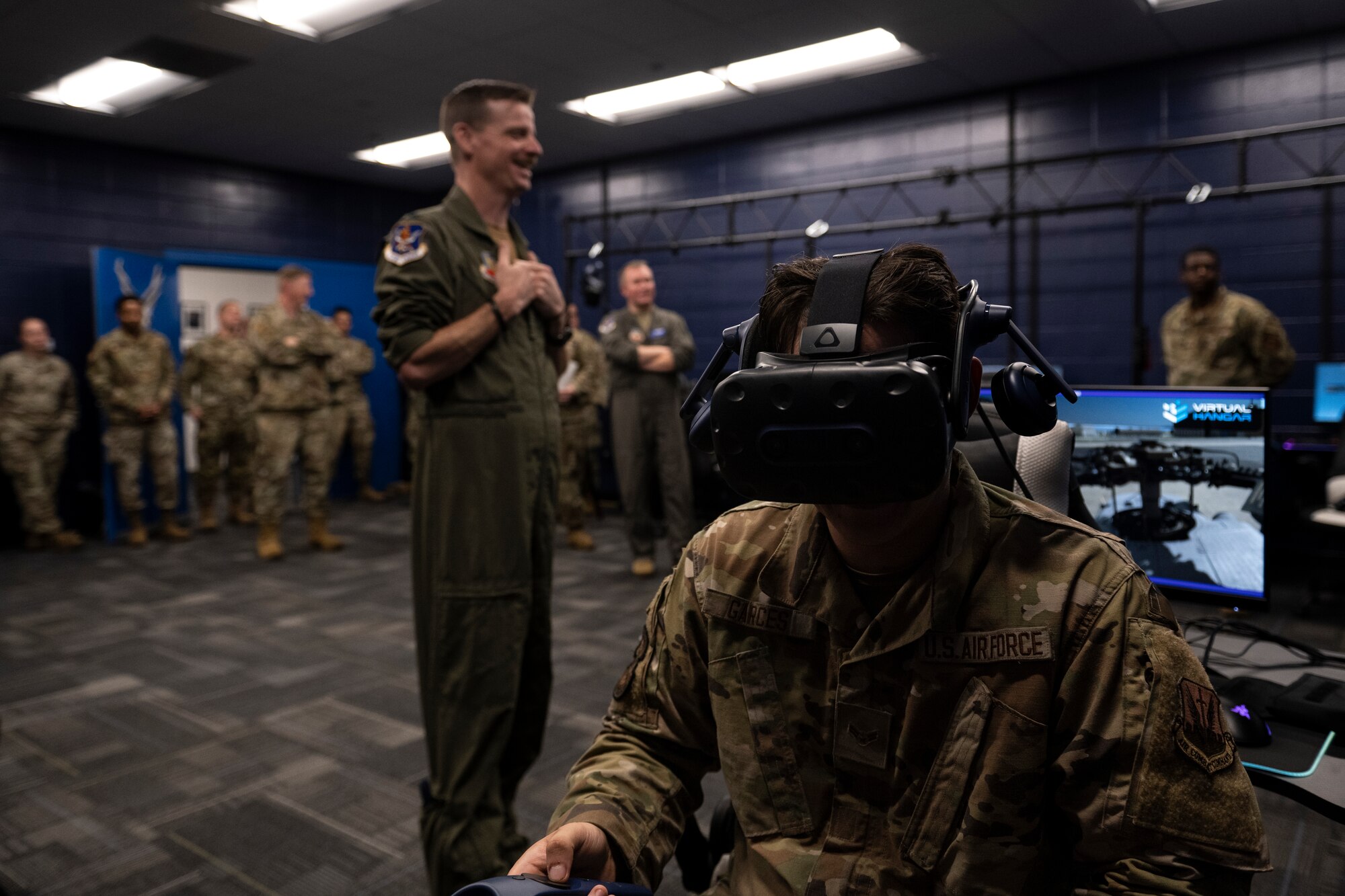 A photo of a ton of Airmen in a room, observing an Airman with a VR headset on.