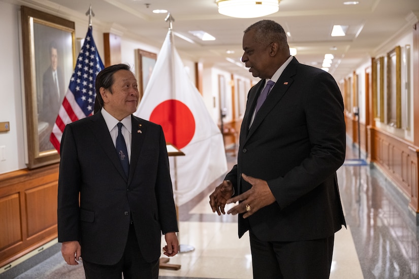 Two men talk in a hallway; U.S. and Japanese flags are in the background.
