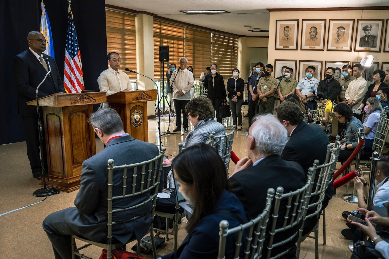 Two men stand behind lecterns as they face a group of people seated in chairs.