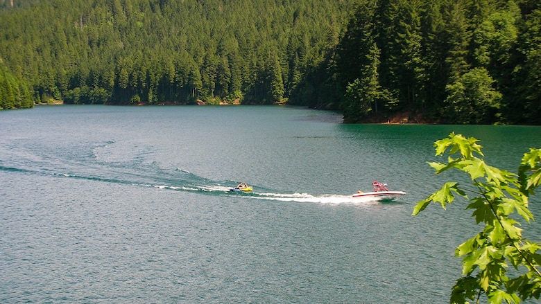 A boat pulling a person on a large tube across a lake surrounded by forest on a sunny day