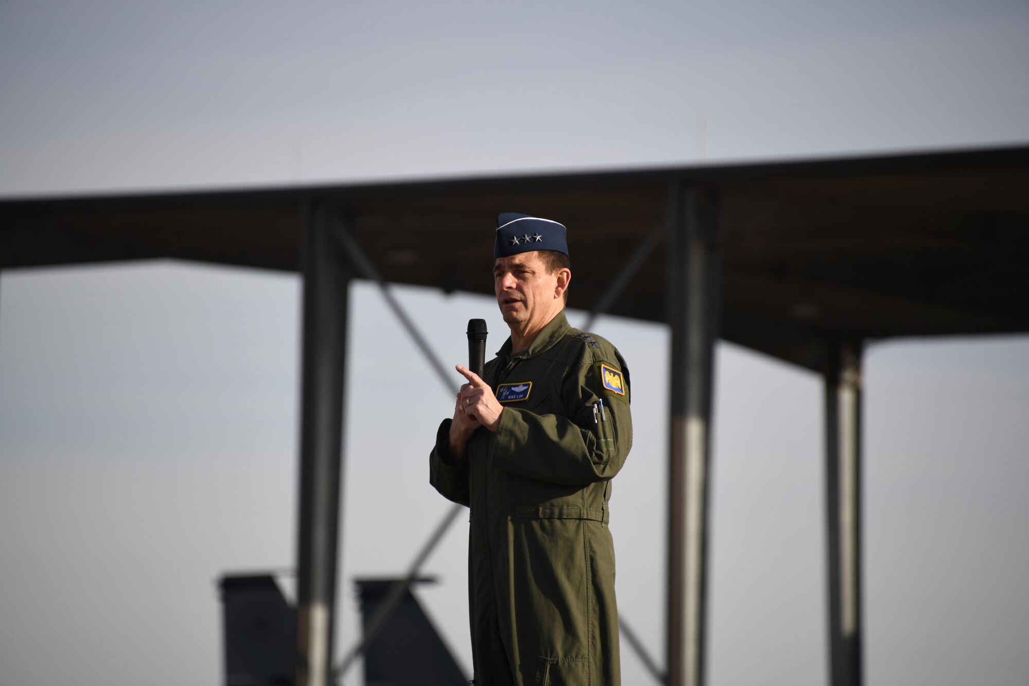 A man wearing a green military flightsuit speaks into a microphone