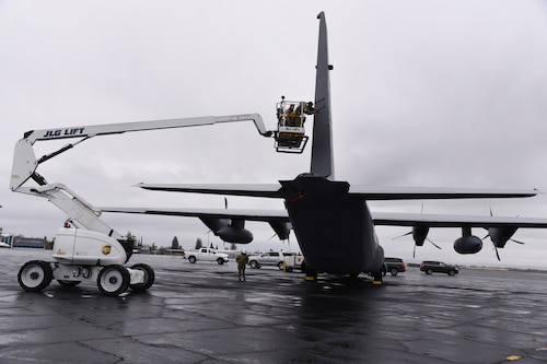 Two Airmen use a lift to reach the tail of the WC-130J aircraft for maintenance, with a safety spotter.