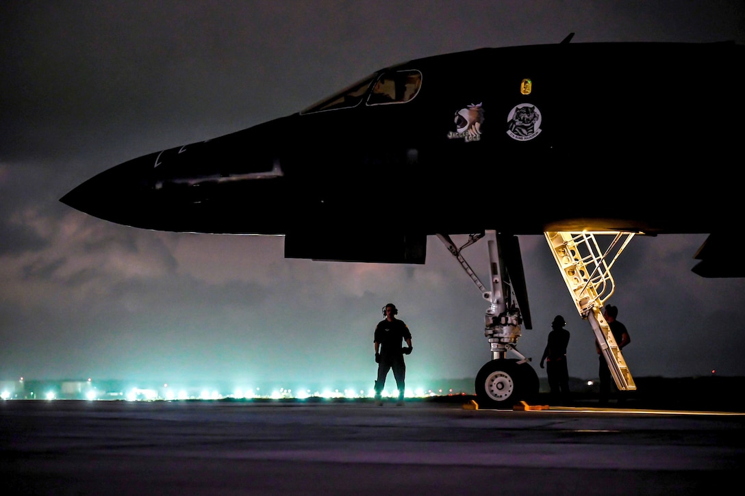 Several airmen stand below a large aircraft lit by runway lights at night.