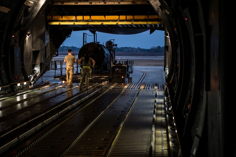 Two airmen watch in an open aircraft as a vehicle moves a large piece of infrastructure into the plane.