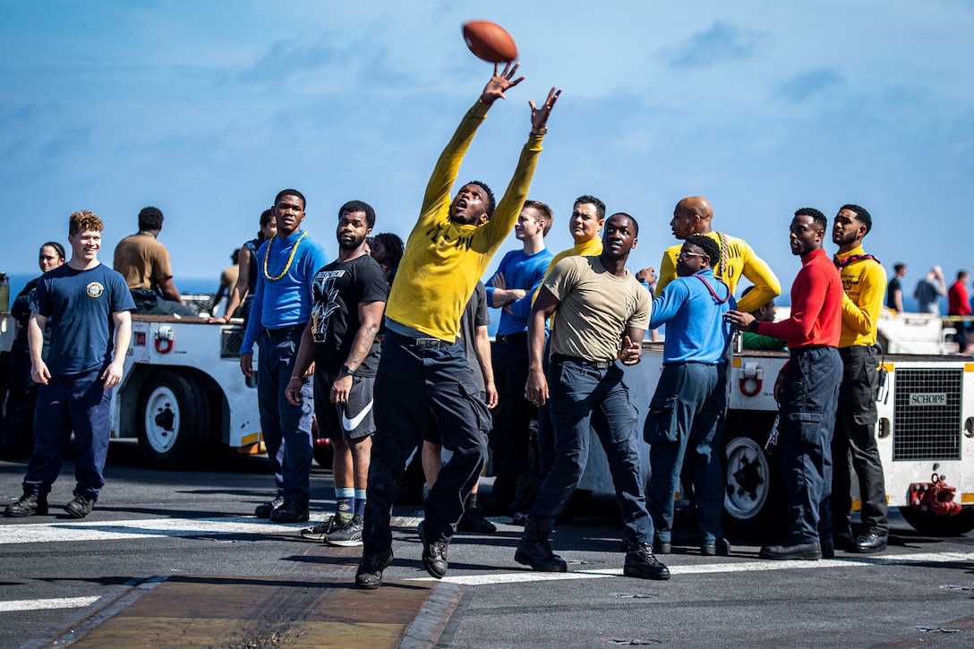 A sailor jumps to reach a football as others look on while standing on the flight deck of a Navy ship.