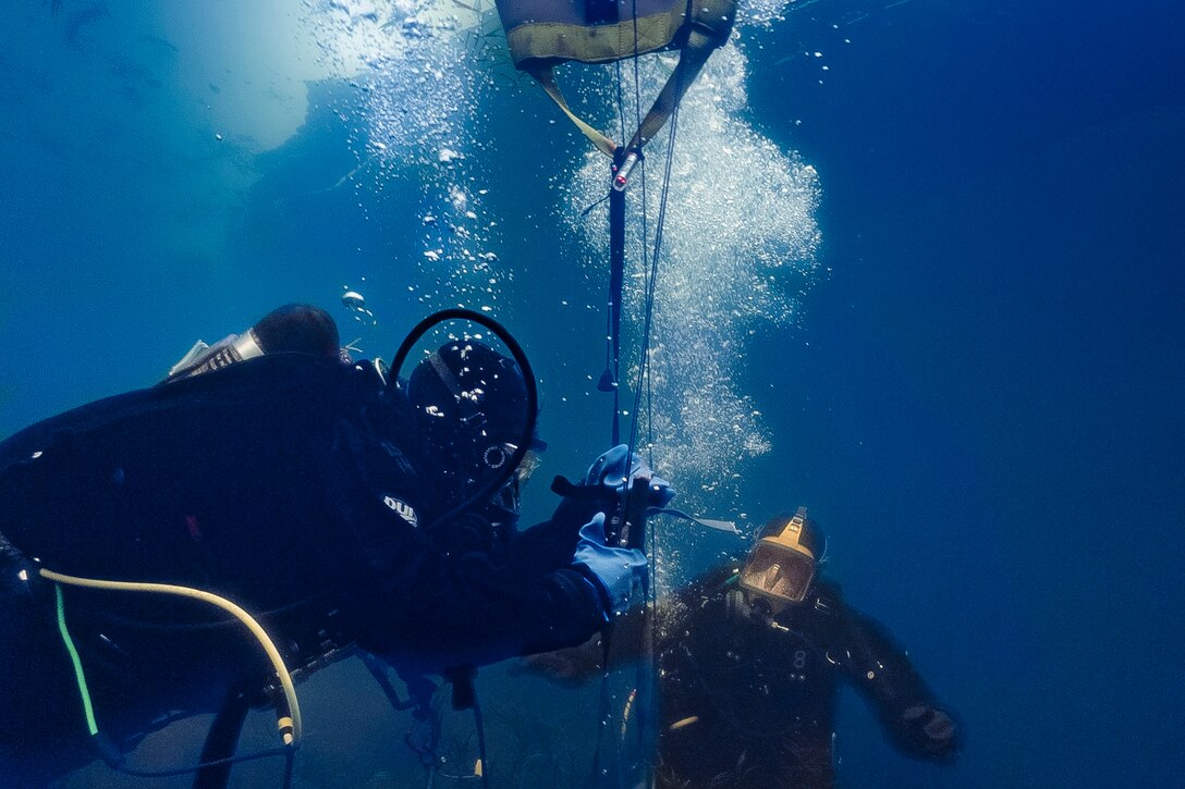 Two sailors in diving gear are shown using a lift bag device underwater.