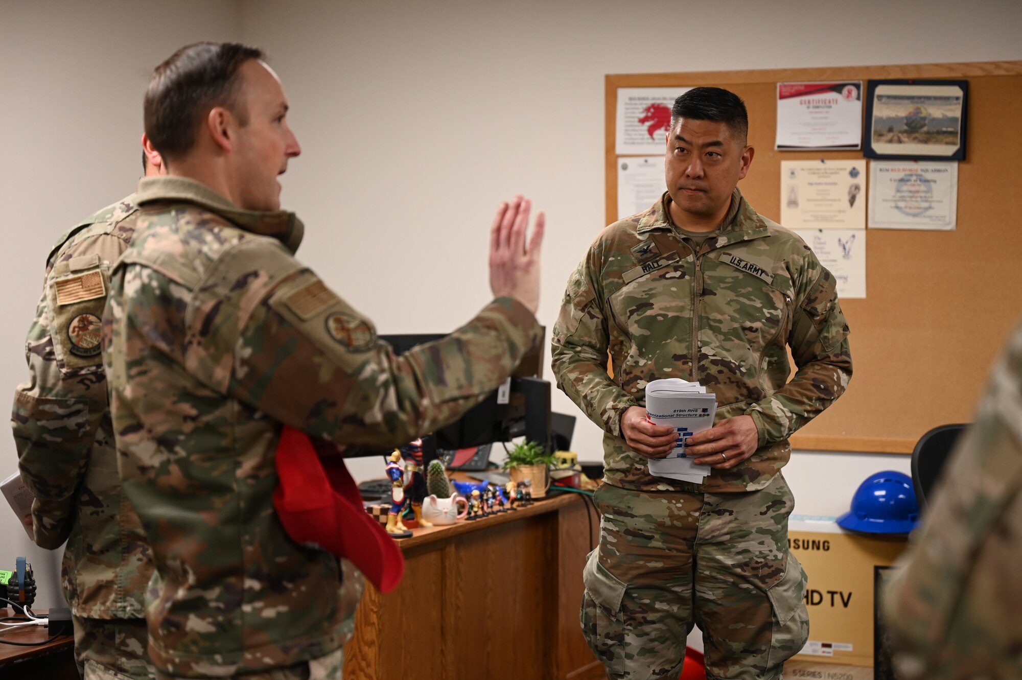 Two men in military uniform stand in an office and talk.