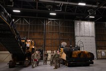 A group of military men stand inside an airfield hangar between two pieces of heavy equipment.