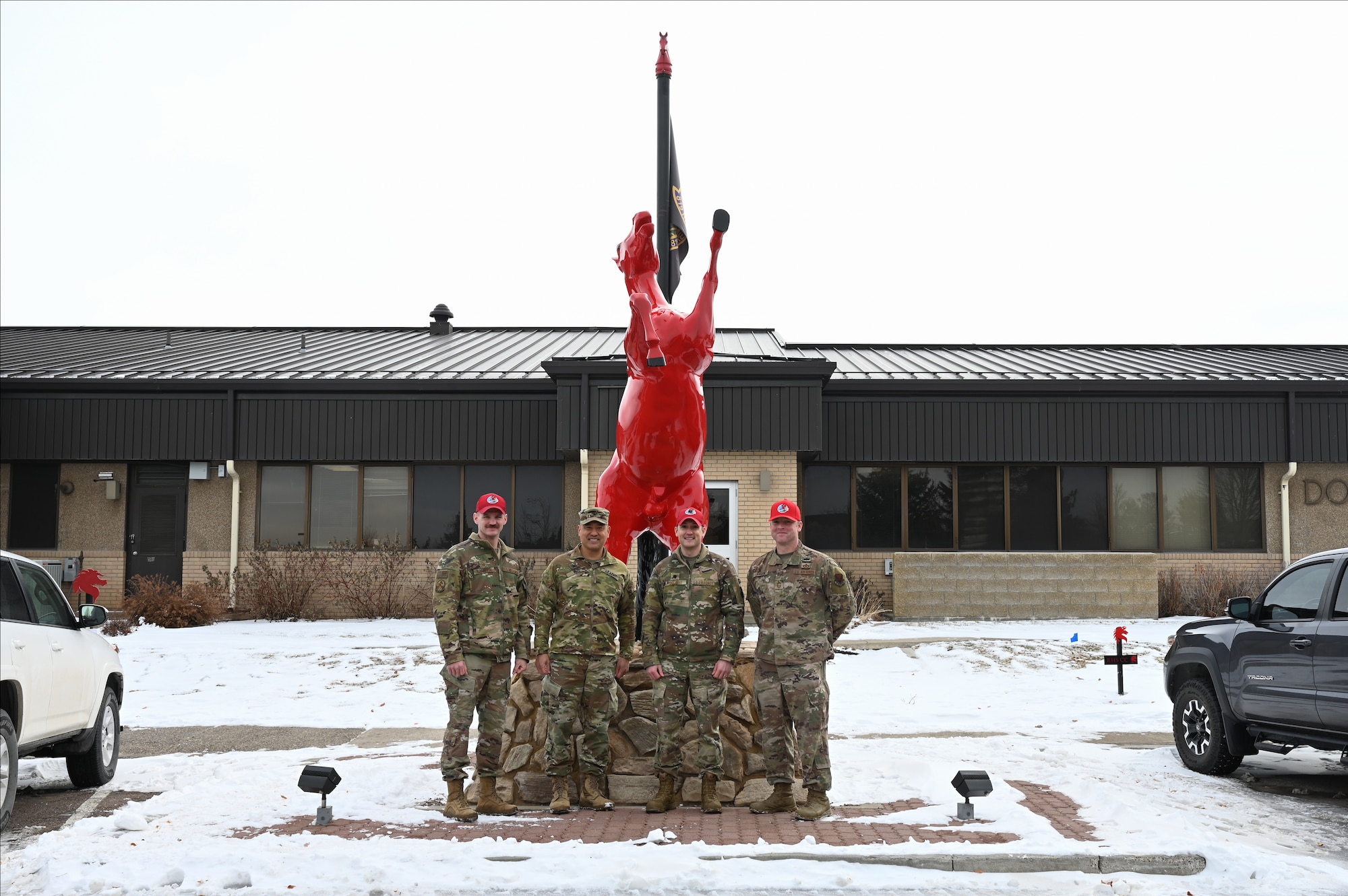 Four uniformed men stand shoulder-to-shoulder for a photo in front of a horse mascot painted red.