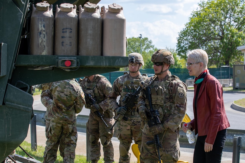 A woman and five male service members look at military equipment.