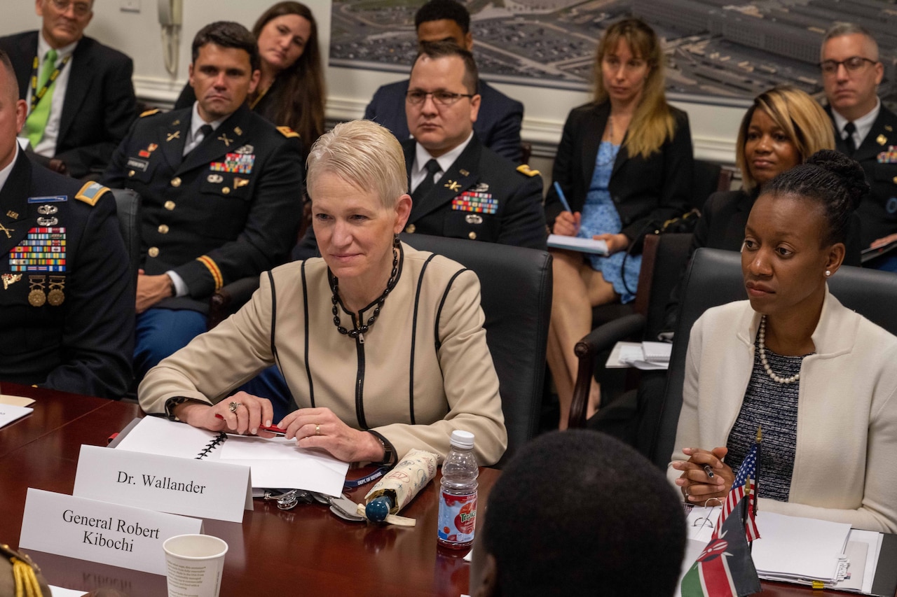 Two women sit at a conference table; others sit in chairs behind them.