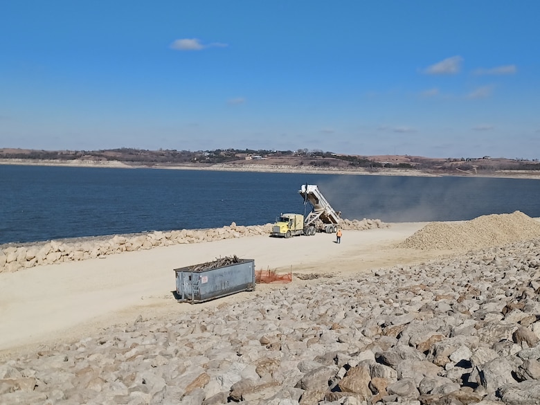 A yellow dump truck dumps large pieces of rock on the ground near a dam with a lake in the background.