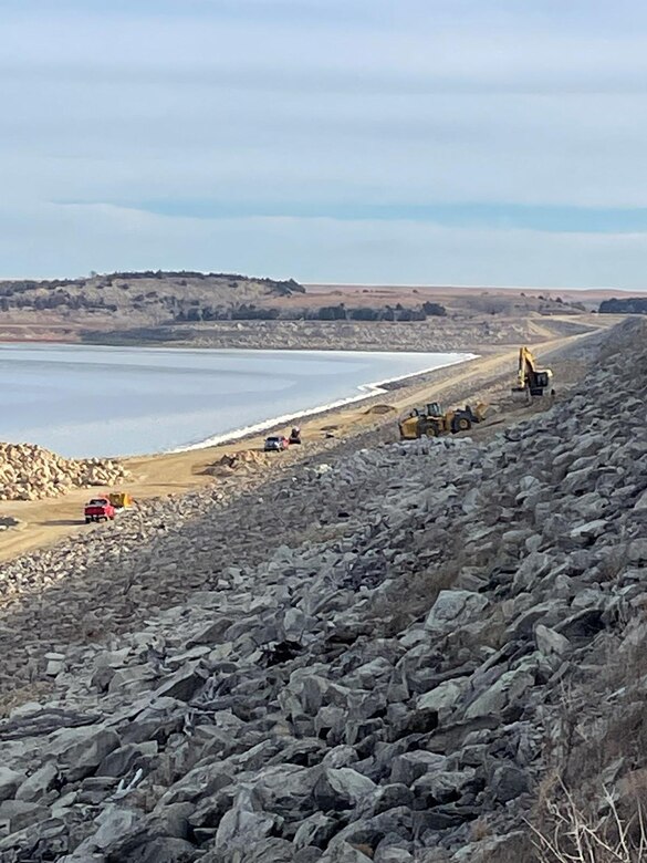 Large machines place large chunks of rock on the face of a dam with a lake to the left and horizon in the back.