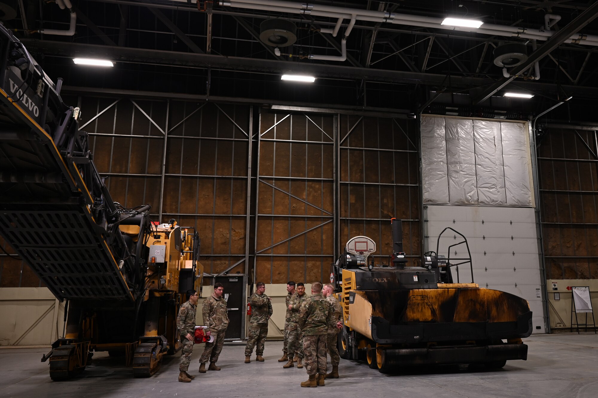 A group of military men stand inside an airfield hangar between two pieces of heavy equipment.