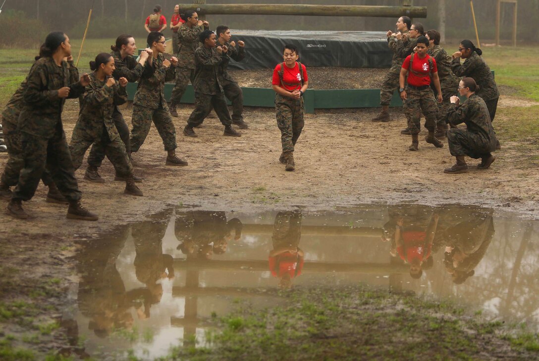A Marine Corps recruit stands in front of a pool of muddy water while others look at her and make fists.